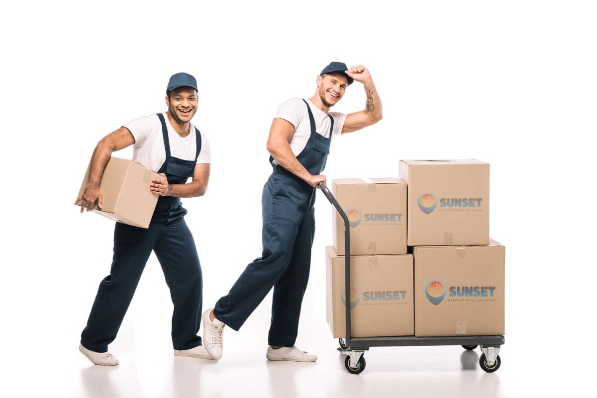 full length of positive multicultural movers in uniform and caps walking near hand truck with carton boxes on white