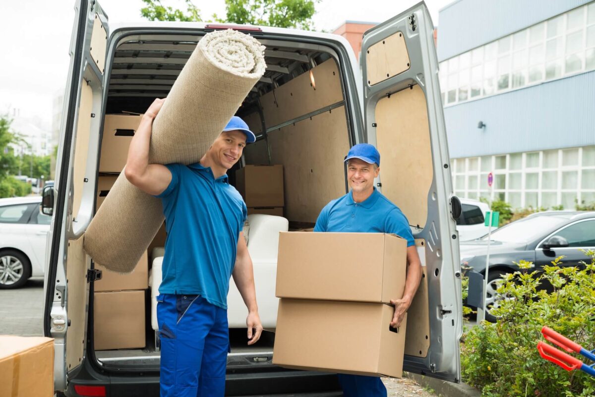 Two movers in front of a van, one holding a carpet, the other holding packages