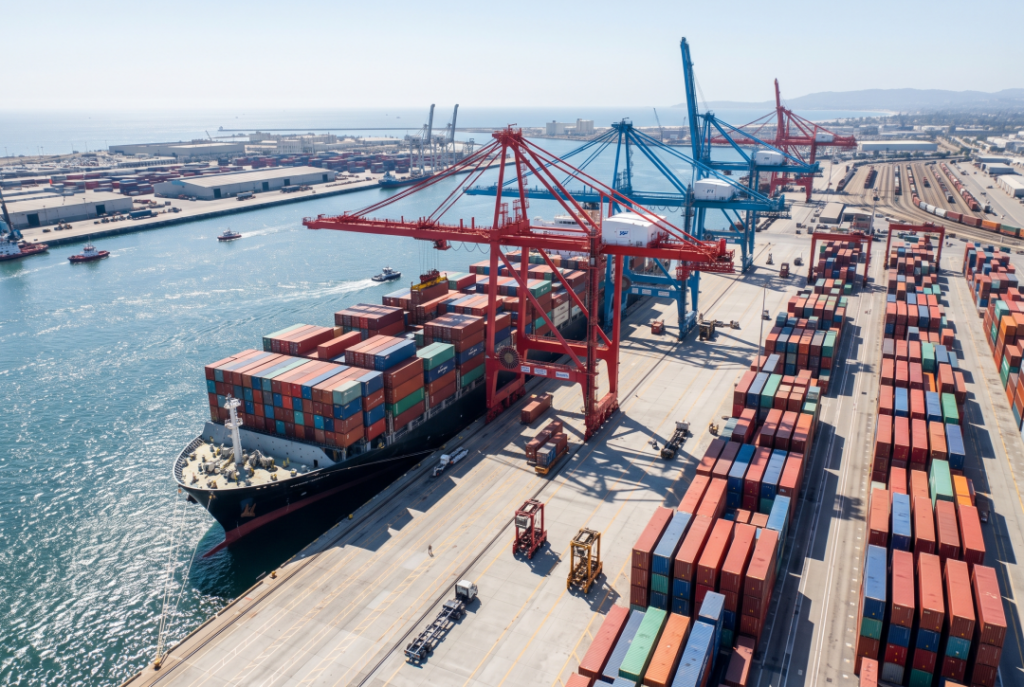 A massive cargo ship is docked at the harbor, with giant industrial cranes carefully loading standard 20-foot and 40-foot steel shipping containers.